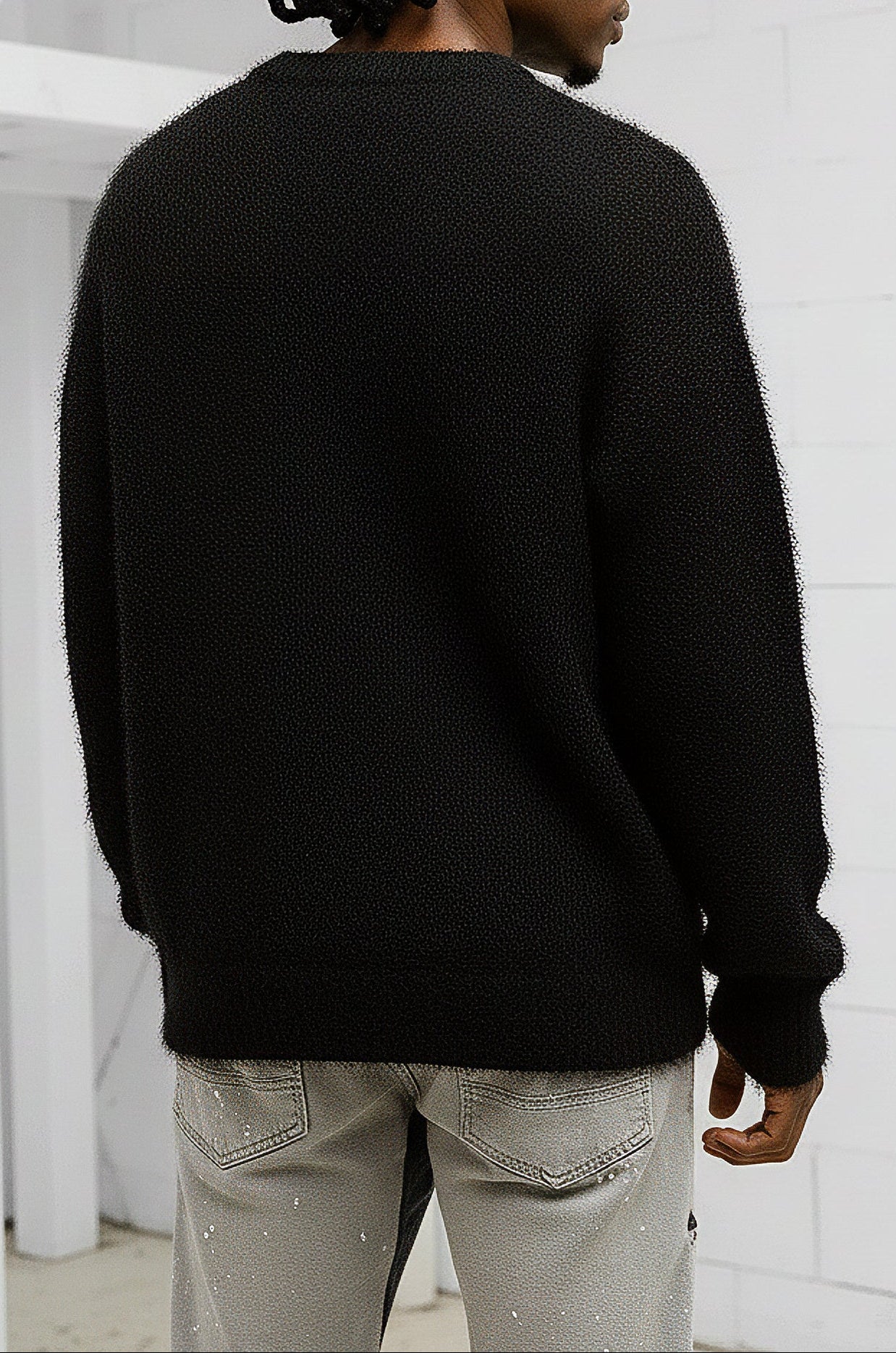 Person wearing a black sweater and light gray pants against a white brick wall.