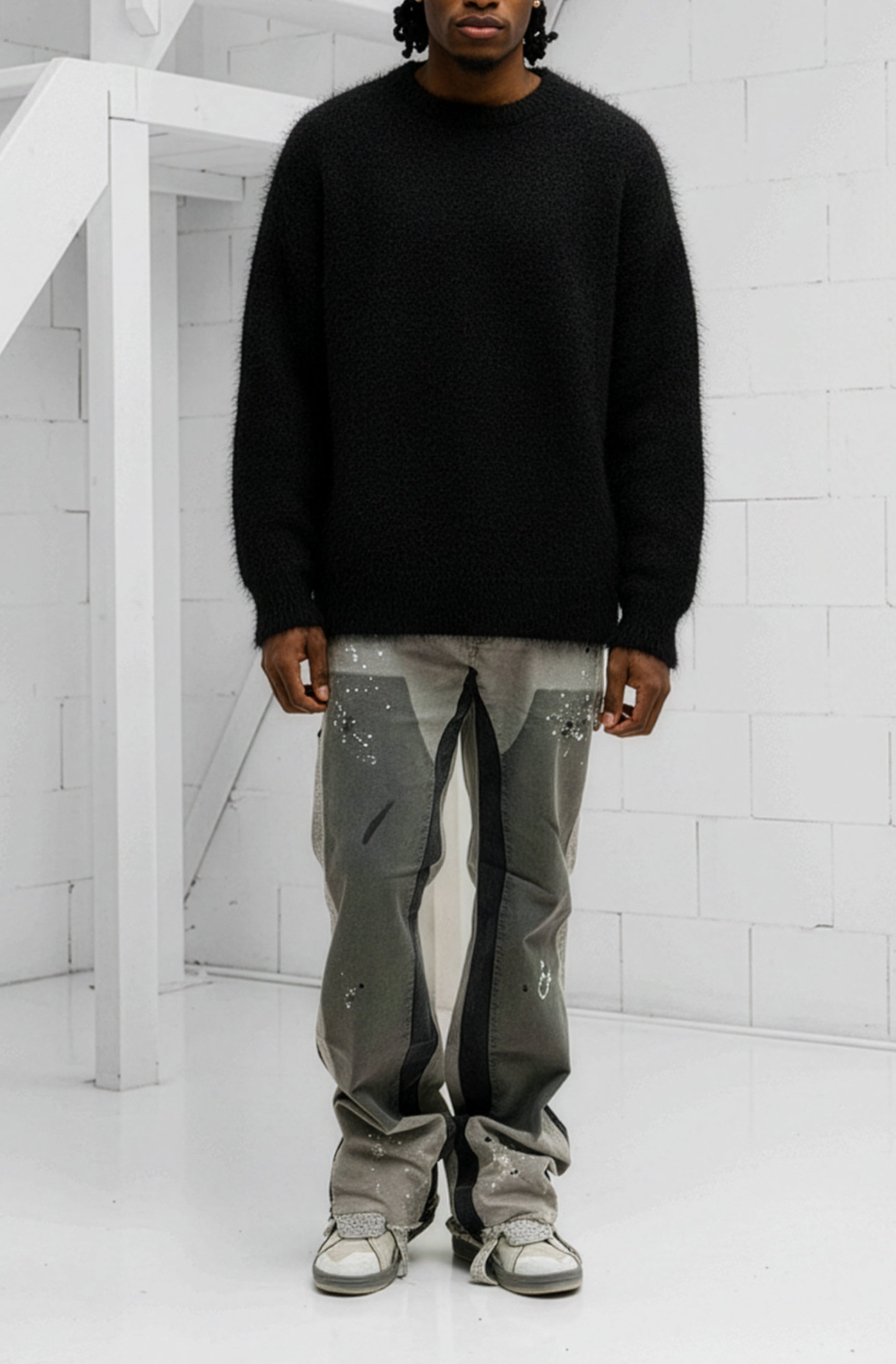 Person wearing a black sweater and gray pants standing in a minimalistic room with white walls and a staircase.
